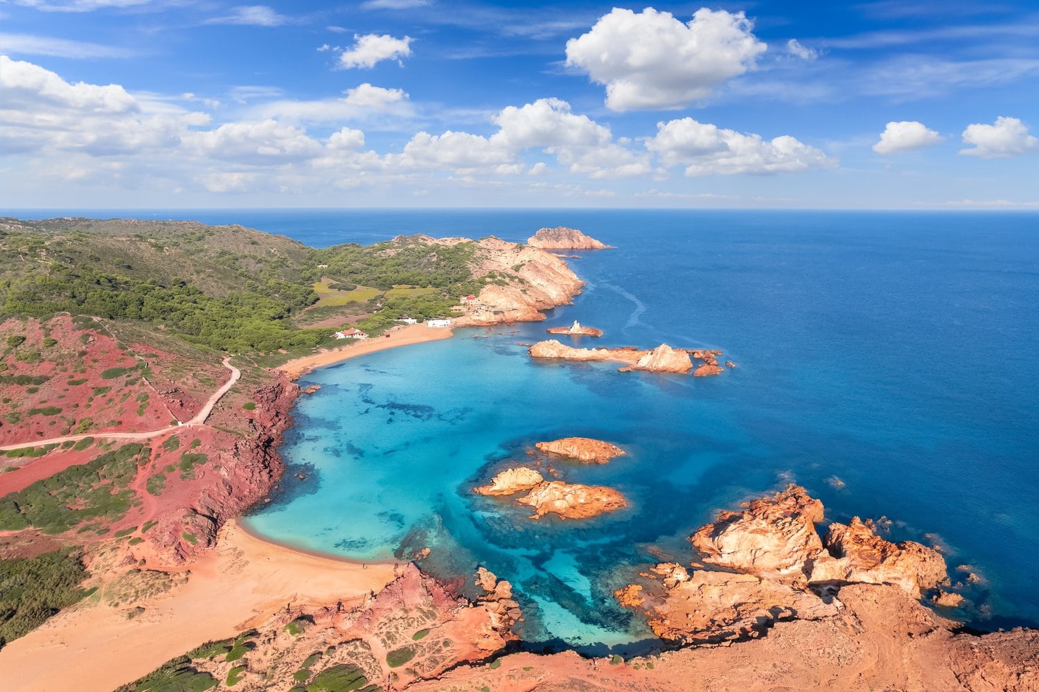 Luftaufnahme der Cala Pregonda im Norden Menorcas mit rostroten Klippen, goldenem Sand und türkisblauem Meer mit vorgelagerten Felsinseln.