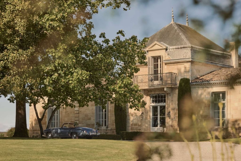 Sandsteinfassade des Château Cordeillan-Bages mit Eckturm und davor parkendem blauen Oldtimer-Cabrio im Schatten alter Bäume.