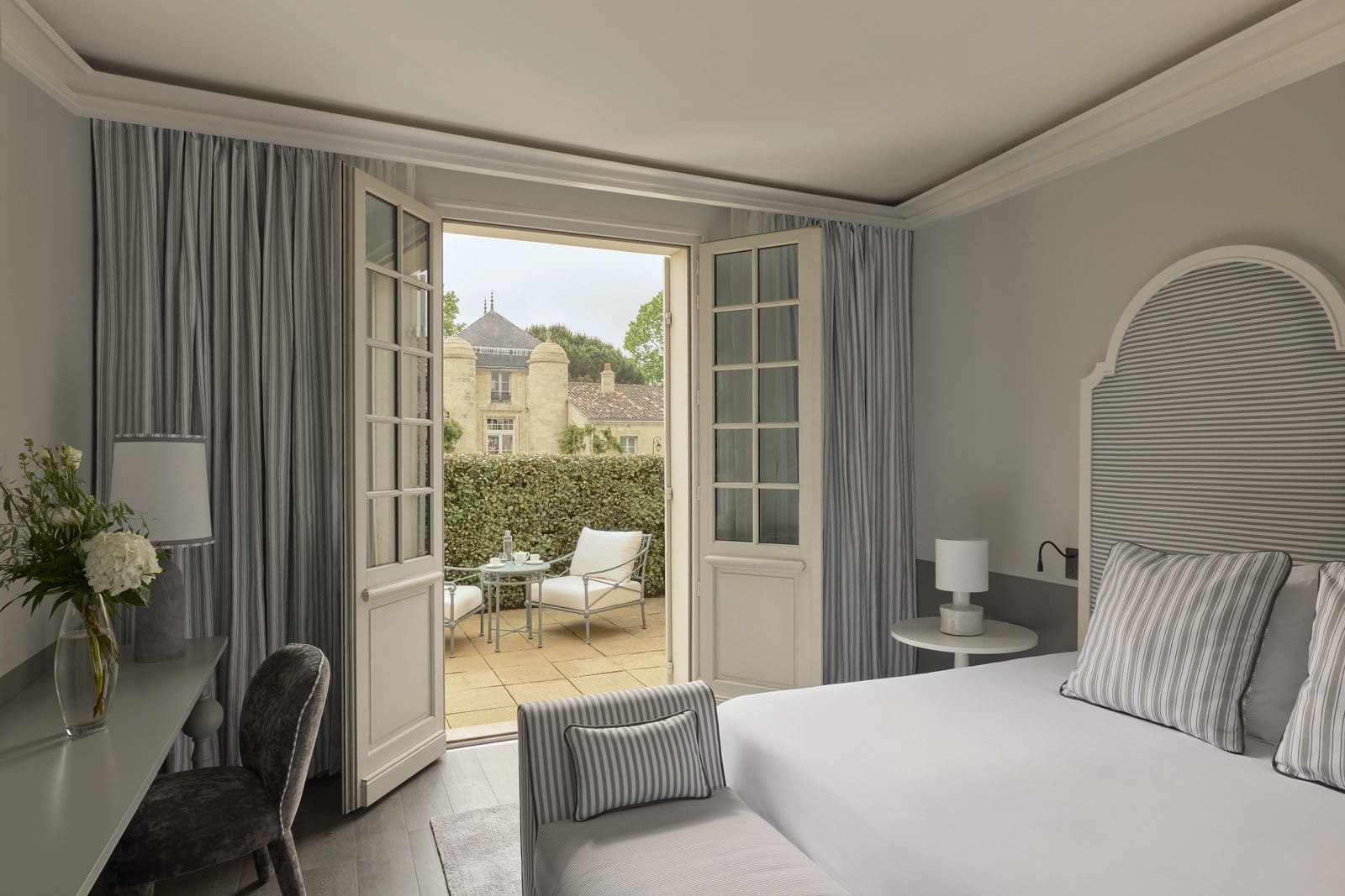 Garden Terrace Room in grauen Tönen mit gestreiften Vorhängen, gepolstertem Bogen-Kopfteil und Blick durch offene Flügeltüren auf die Privatterrasse.