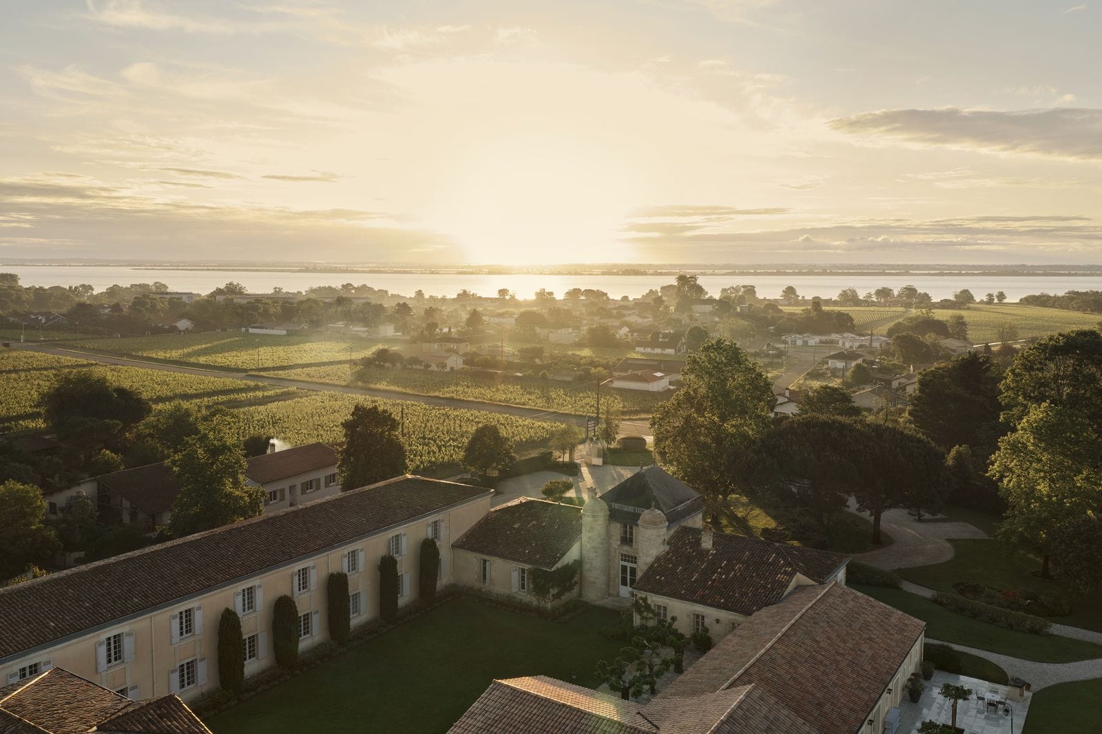 Luftaufnahme von Château Cordeillan-Bages bei Sonnenuntergang mit umliegenden Weinbergen und der Gironde-Mündung im Hintergrund.