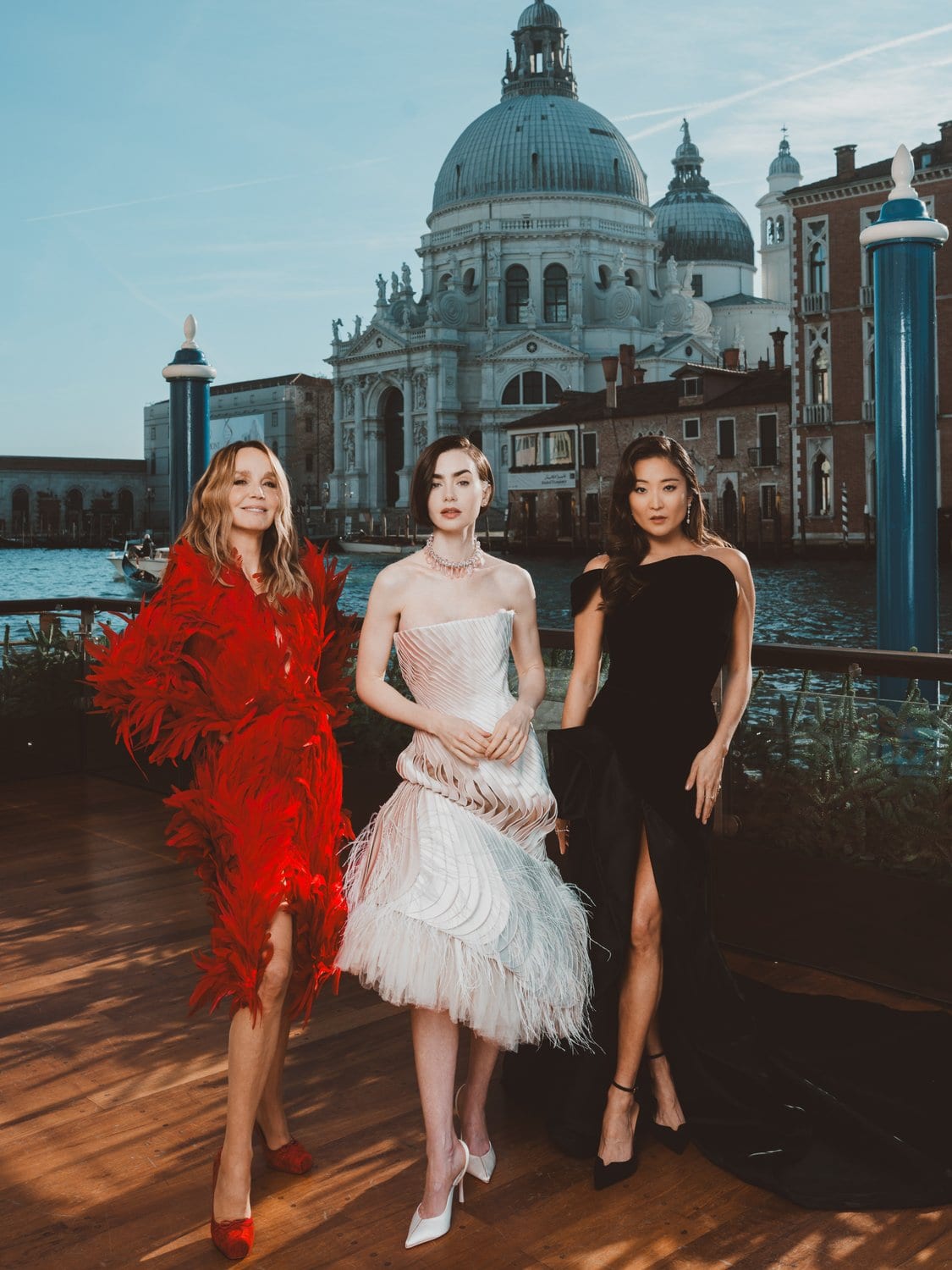 Der weibliche Cast von Emily in Paris - Drei Frauen in glamourösen Abendkleidern posieren auf einem Holzsteg am Canal Grande in Venedig, im Hintergrund die Kuppel der Basilika Santa Maria della Salute im Abendlicht