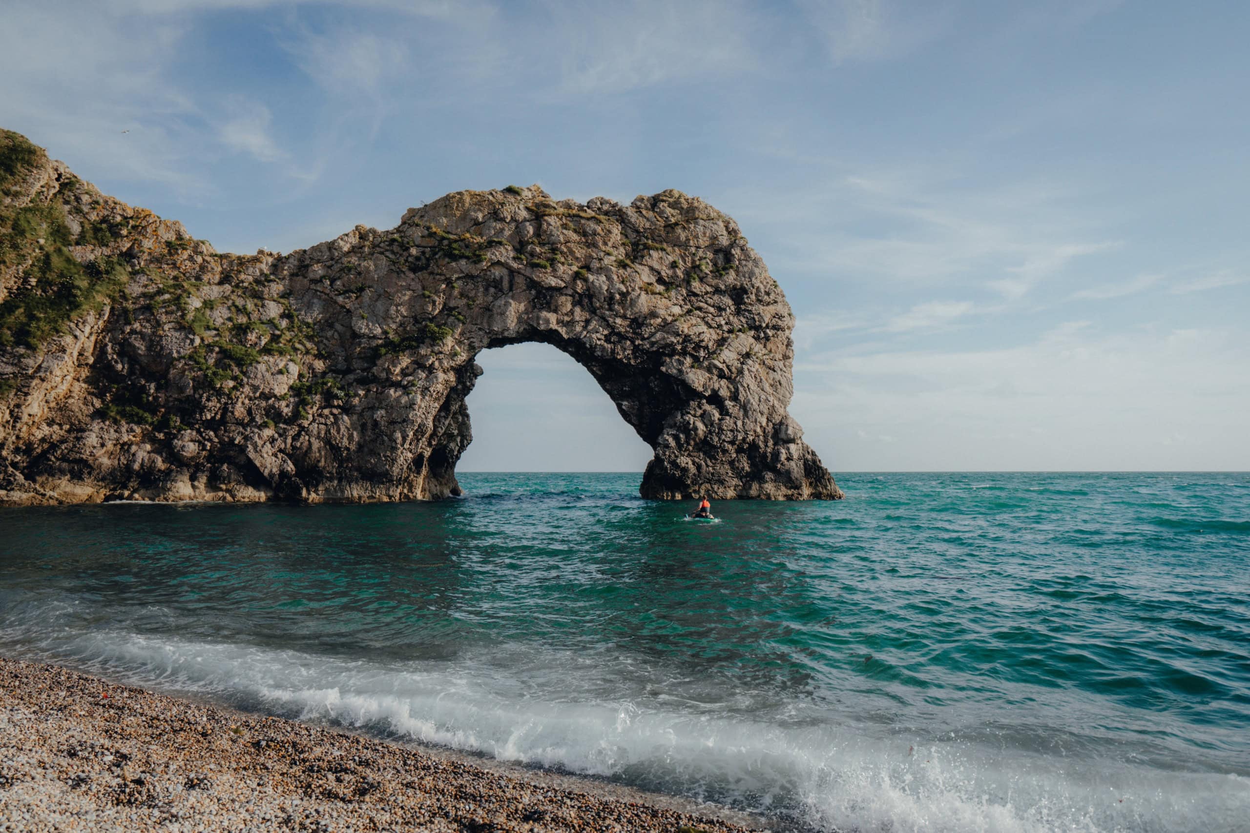 Ein Felsvorsprung reicht in die Meeresbrandung an einem Strand hinein. Im Vorsprung selbst bildet eine Auslassung eine Art rundes Tor: die Durdle Door.