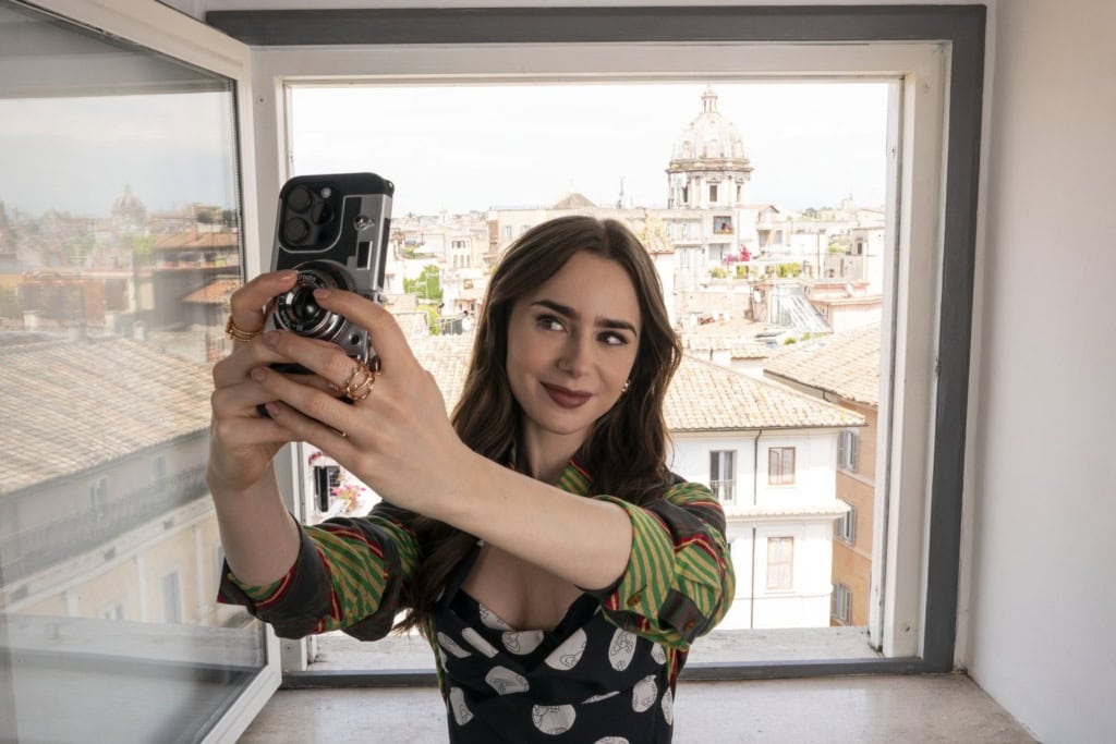 Junge Frau mit langen braunen Haaren macht ein Selfie mit Smartphone und kompakter Filmkamera am offenen Fenster, im Hintergrund die Dächer und eine Kirchenkuppel von Rom
