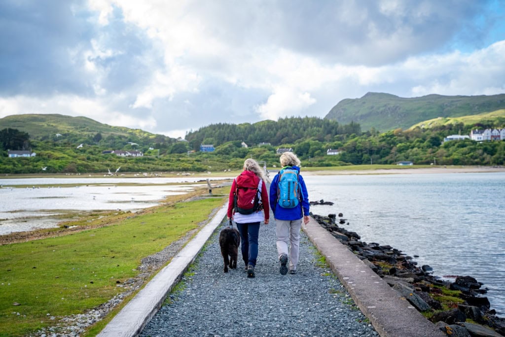 Ein von Stein umfasster grüner Pfad führt geradwegs über eine Meerenge auf eine Insel. Darauf wandern zwei Menschen in Outdoorkleidung. Im Hintergrund sind grüne Berge erkennbar.