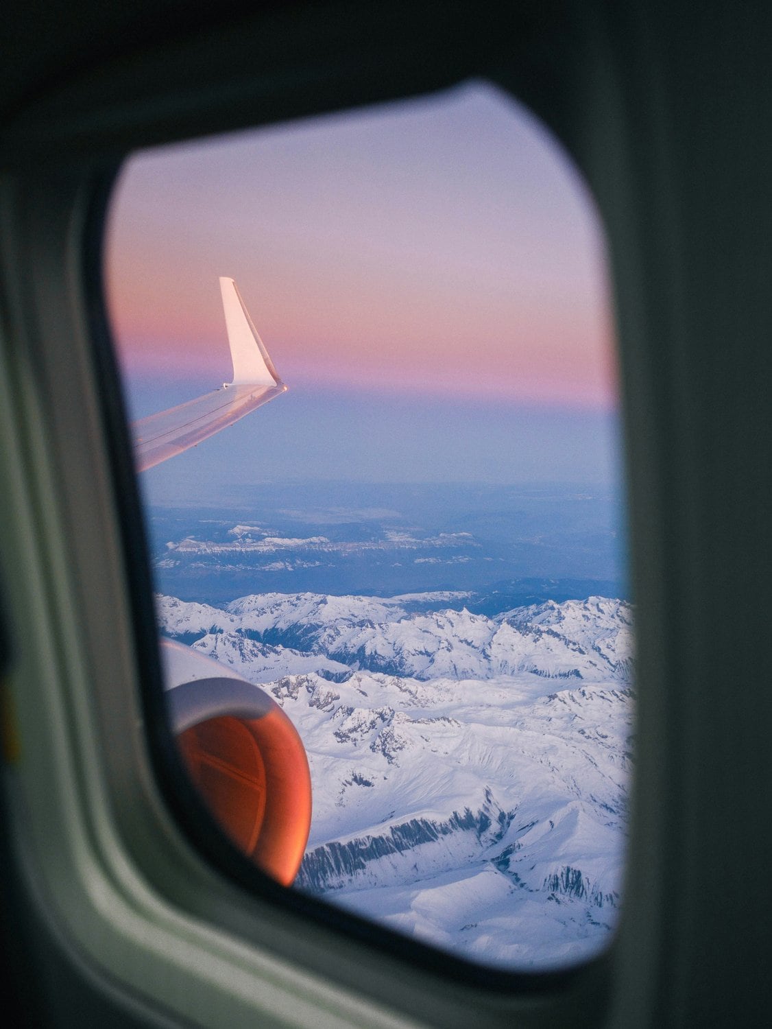 Blick aus einem Flugzeugfenster auf Tragfläche, Triebwerk und schneebedeckte Alpen im rosafarbenen Sonnenuntergangslicht