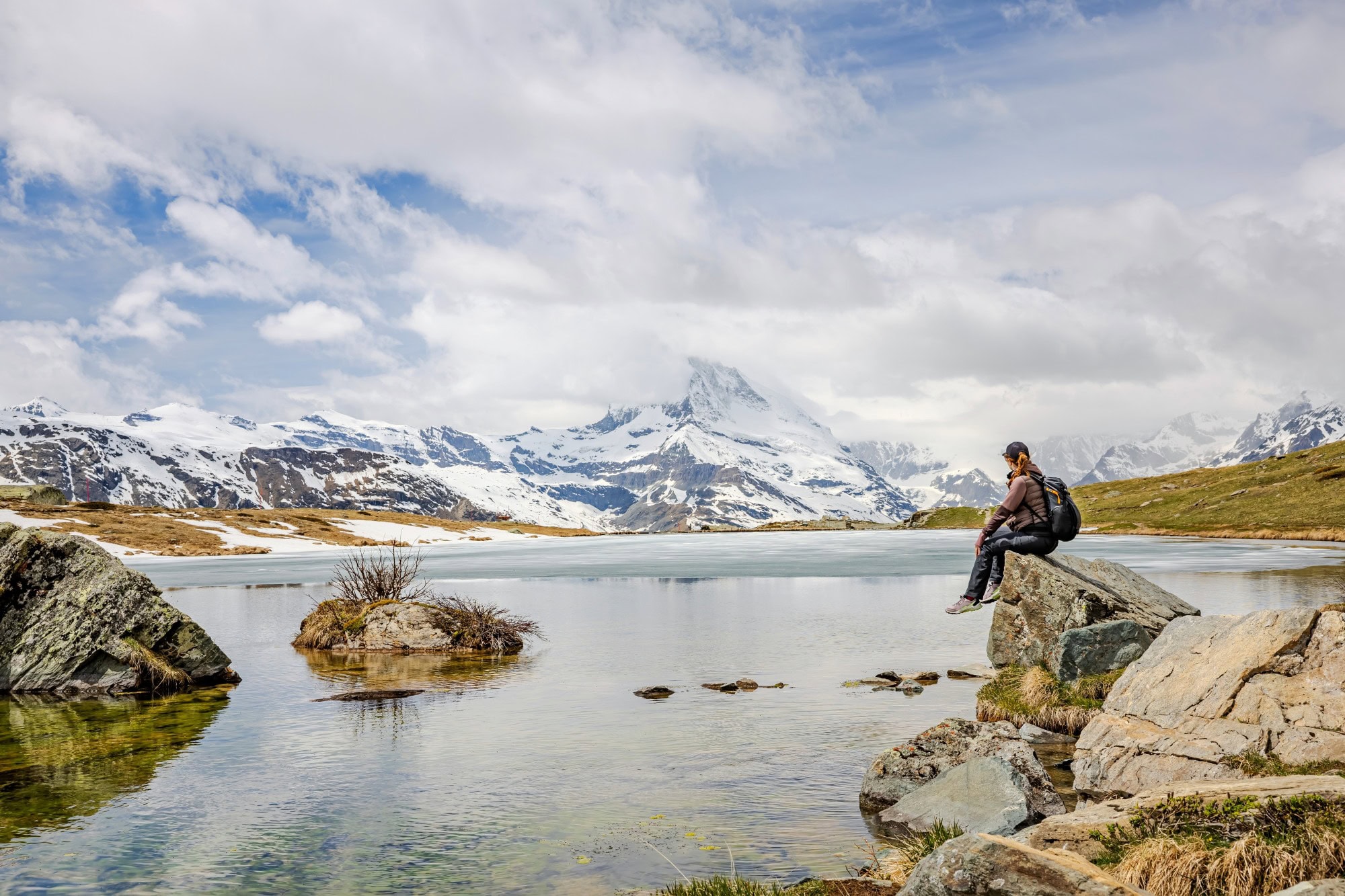 Eine Frau sitzt am Ufer eines großen Bergsees auf einem Felsvorsprung und schaut Richtung Horizont, wo am anderen Ufer des Sees ein riesiger, schneebdeckter Berg in die Wolken hineinragt.