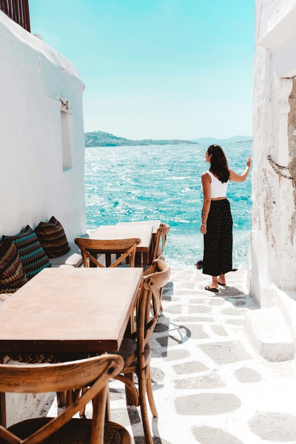 Frau in weißem Top und schwarzem Boho-Rock steht in einer engen weißgetünchten Gasse und blickt auf das tiefblaue Ägäische Meer, im Vordergrund Holztische einer Taverne