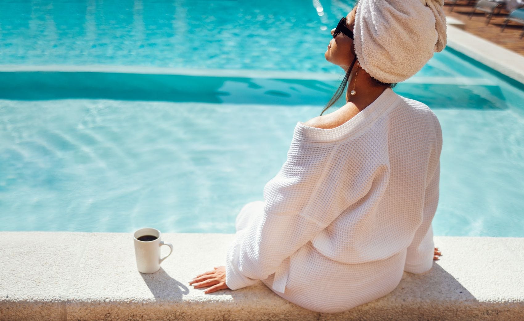 Junge Frau genießt am Hotelpool die Sonne, neben ihr eine Tasse Kaffee 