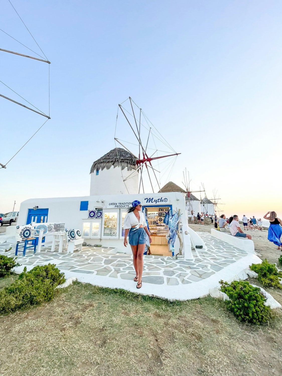 Junge Frau in Jeansshorts und weißem Hemd steht im Abendlicht vor einer traditionellen Mykonos-Windmühle, daneben der kleine Souvenirshop „Mytho"