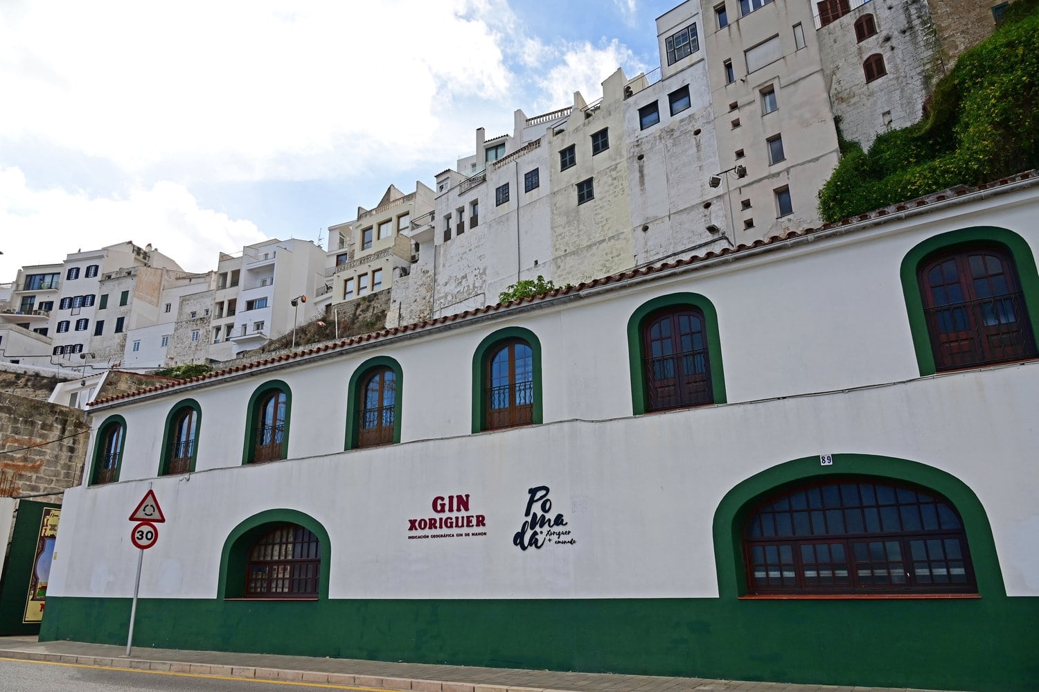 Fassade der Gin-Xoriguer-Destillerie am Hafen von Mahón mit grün gerahmten Bogenfenstern, darüber die weißen Häuser der Altstadt am Felshang