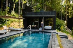 Ein moderner, in graue Betonplatten eingelassener Pool liegt in einem Wald. Am linken Rand zwei helle Liegen, vor Kopf eine hölzerne Sauna, vor der eine Frau in Bademantel und mit Tasche entlang läuft. Der Pool scheint inmitten eines Walds zu liegen.