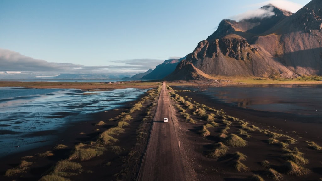 Drohnenaufnahme eines weißen Autos, das über eine langezogene Schotterpiste auf Island Richtung Horizont fährt, wo sich zur rechten ein gewaltiges Gebirge auftürmt.