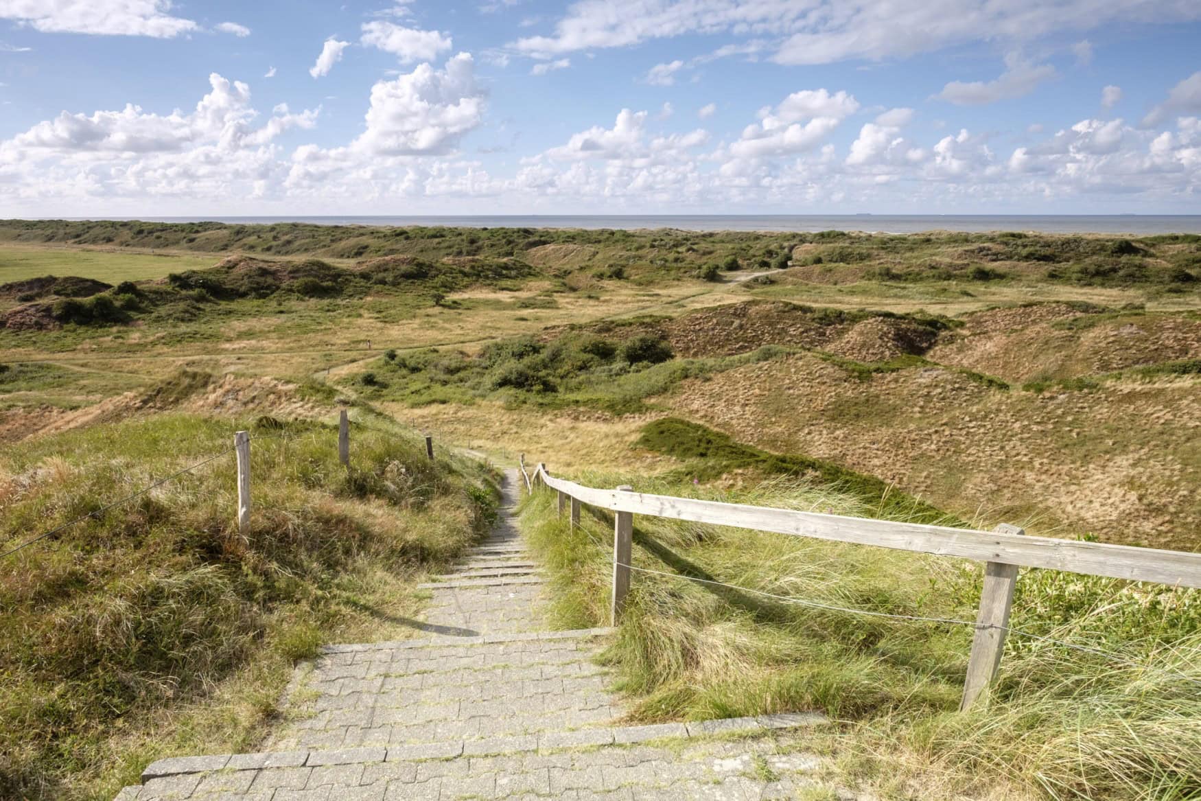 Vom Scheitel einer Düne führt eine geziegelte Treppe hinab ins Dünental. Im Hintergrund ist die Nordsee erkennbar.