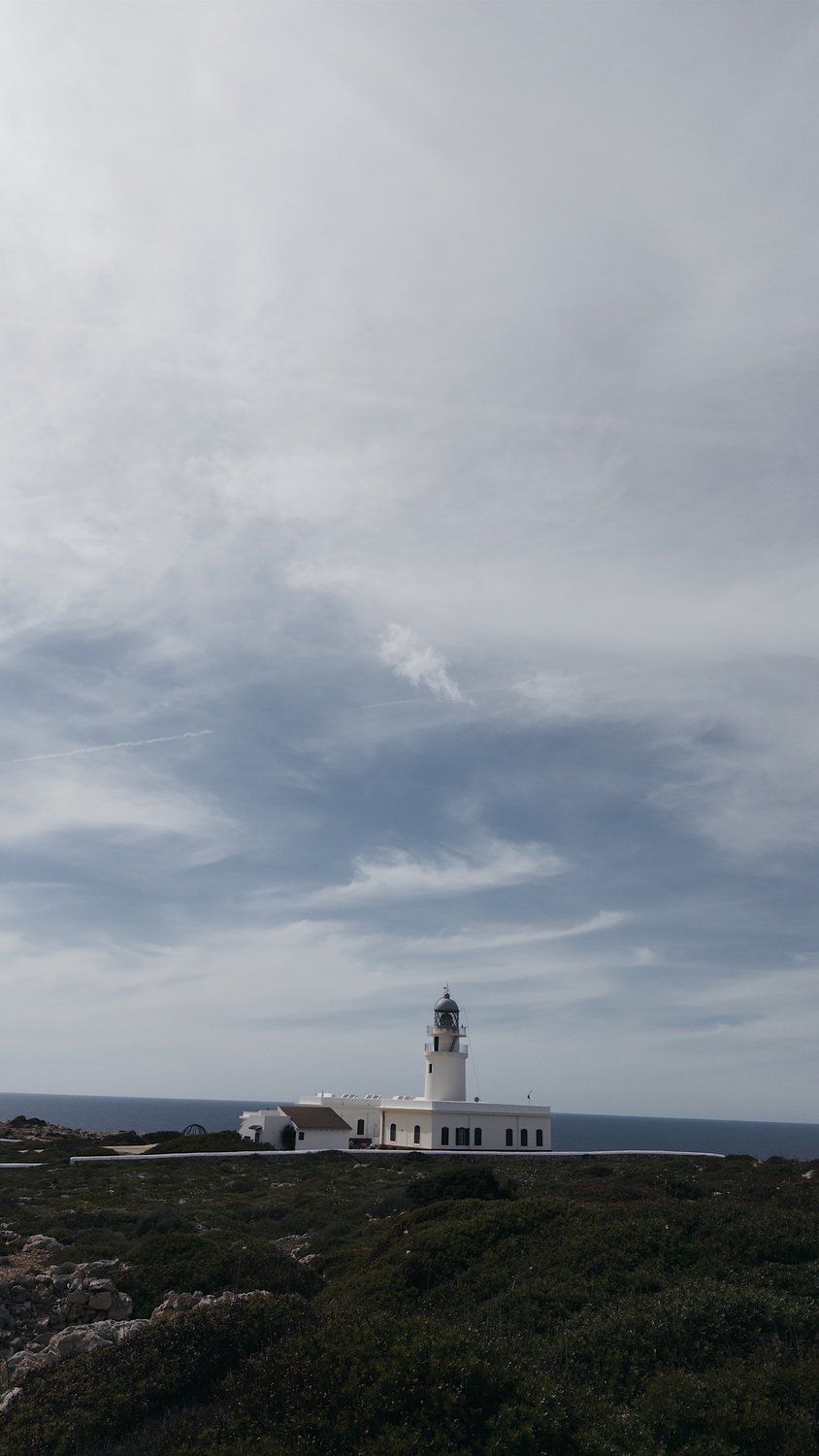 Weißer Leuchtturm Far de Cavalleria auf einer kahlen Klippe im Norden Menorcas vor weitem Himmel mit Schleierwolken