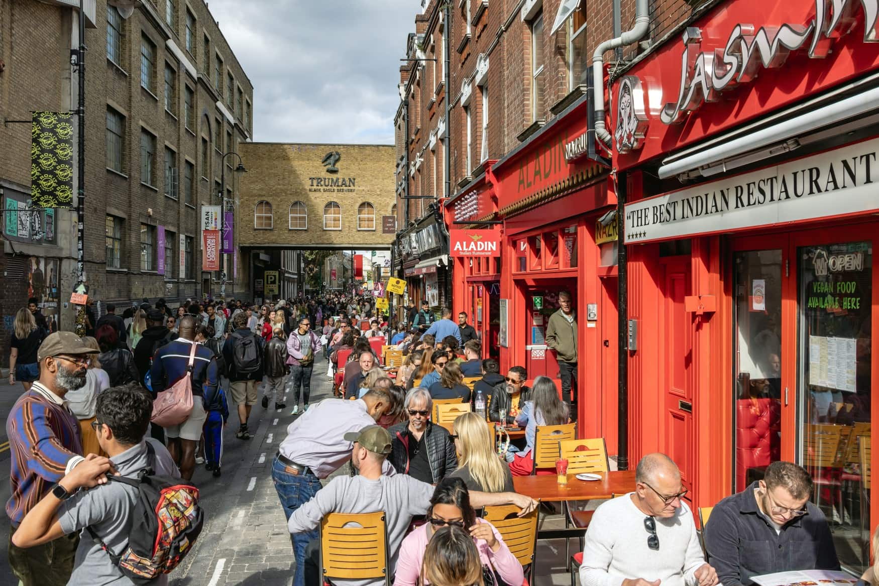 Eine Seitenstraße der Brick Lane in London im Sommer; vor mehreren Restaurants sind die Tische gut gefüllt.