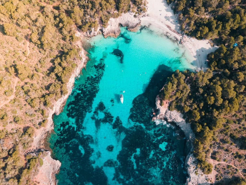 Senkrechte Drohnenaufnahme der Cala en Turqueta mit gestochen klarem türkisem Wasser, weißem Sandstrand, einem ankernden Segelboot und dichtem Pinienwald.
