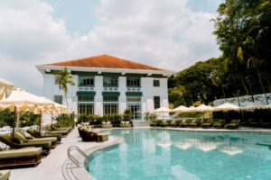 Vor einem Hotel in weißem Kolonialstil in grüner Umgebung liegt ein geschwungener Pool mit Liegen an beiden Seiten.