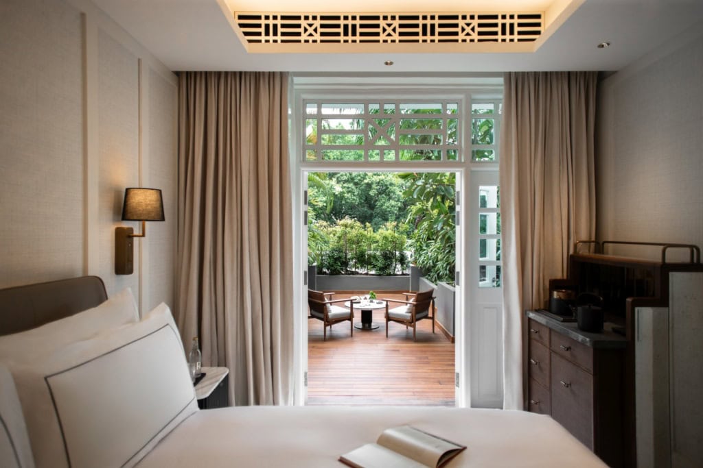 Hinter einem breiten Hotelbett liegt eine Doppeltür, die den Weg zu einer parkettgedeckten Terrasse öffnet. Die Terrasse selbst ist umgeben von tropischer Begrünung.