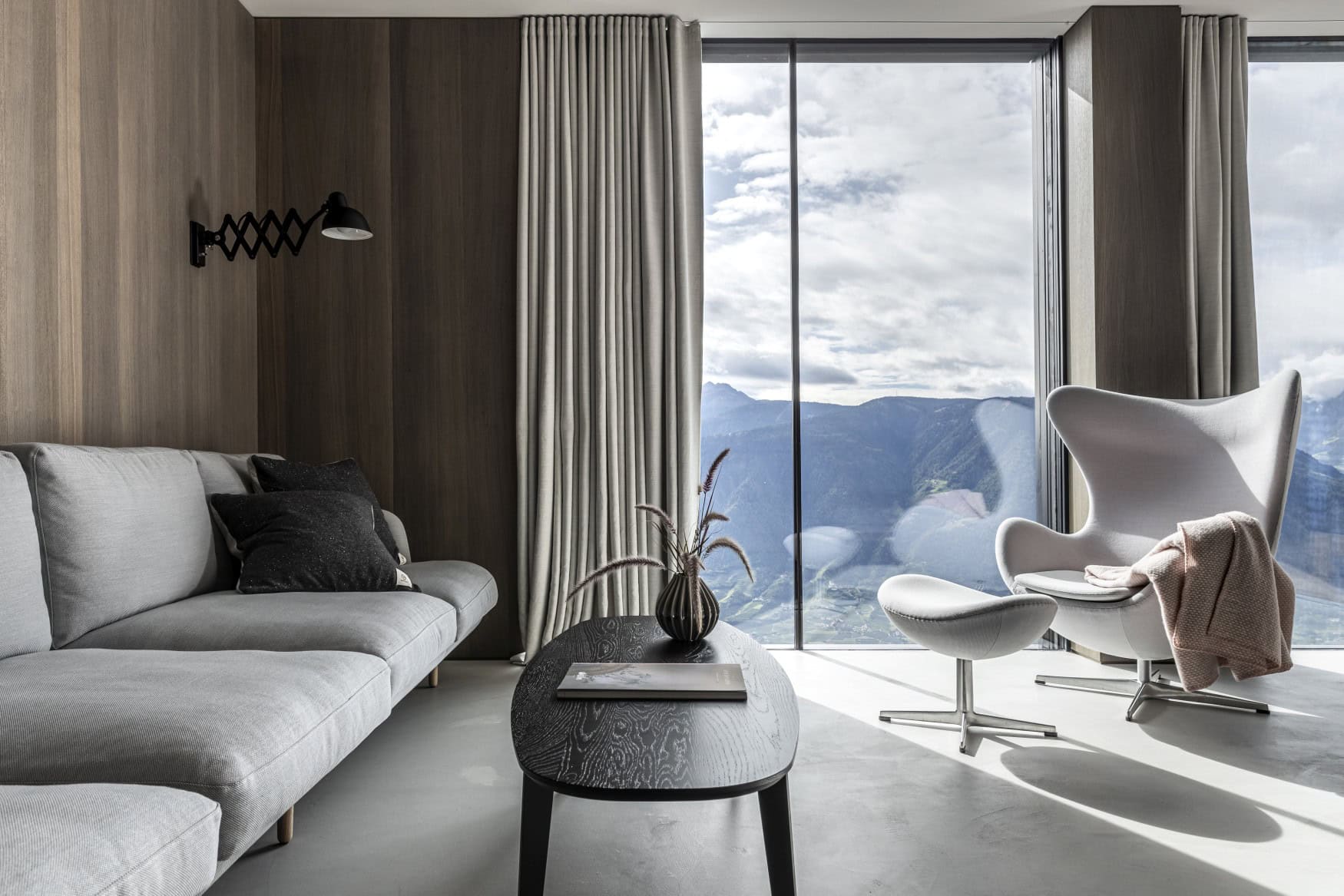 Eine Sitzecke mit grauer Polstercouch und grauem Polstersessel vor einer Glaswand, die den Blick auf die Alpen freigibt.