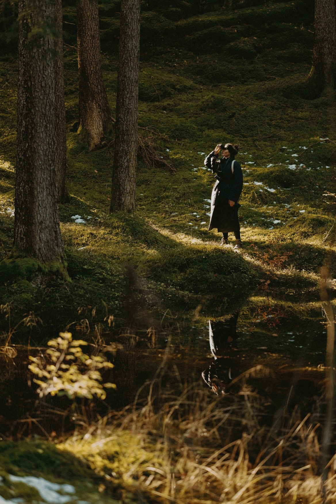 In einem grünen Waldstück steht eine junge Frau in schwarzem Daunenmantel und schaut durch ein Fernglas.
