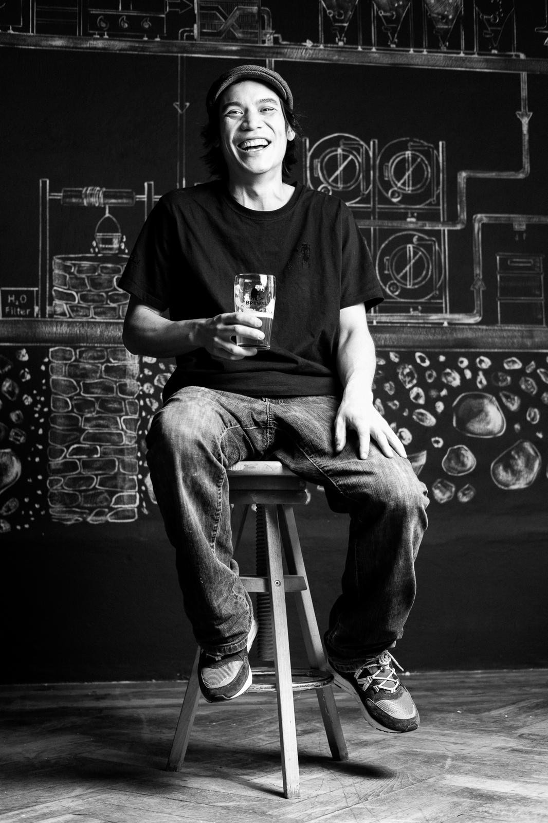 Schwarz-Weiß-Foto eines jungen Mannes, der in Jeans und T-Shirt mit einem Bier in der Hand auf einem Barhocker sitzt und in die Kamera lacht.