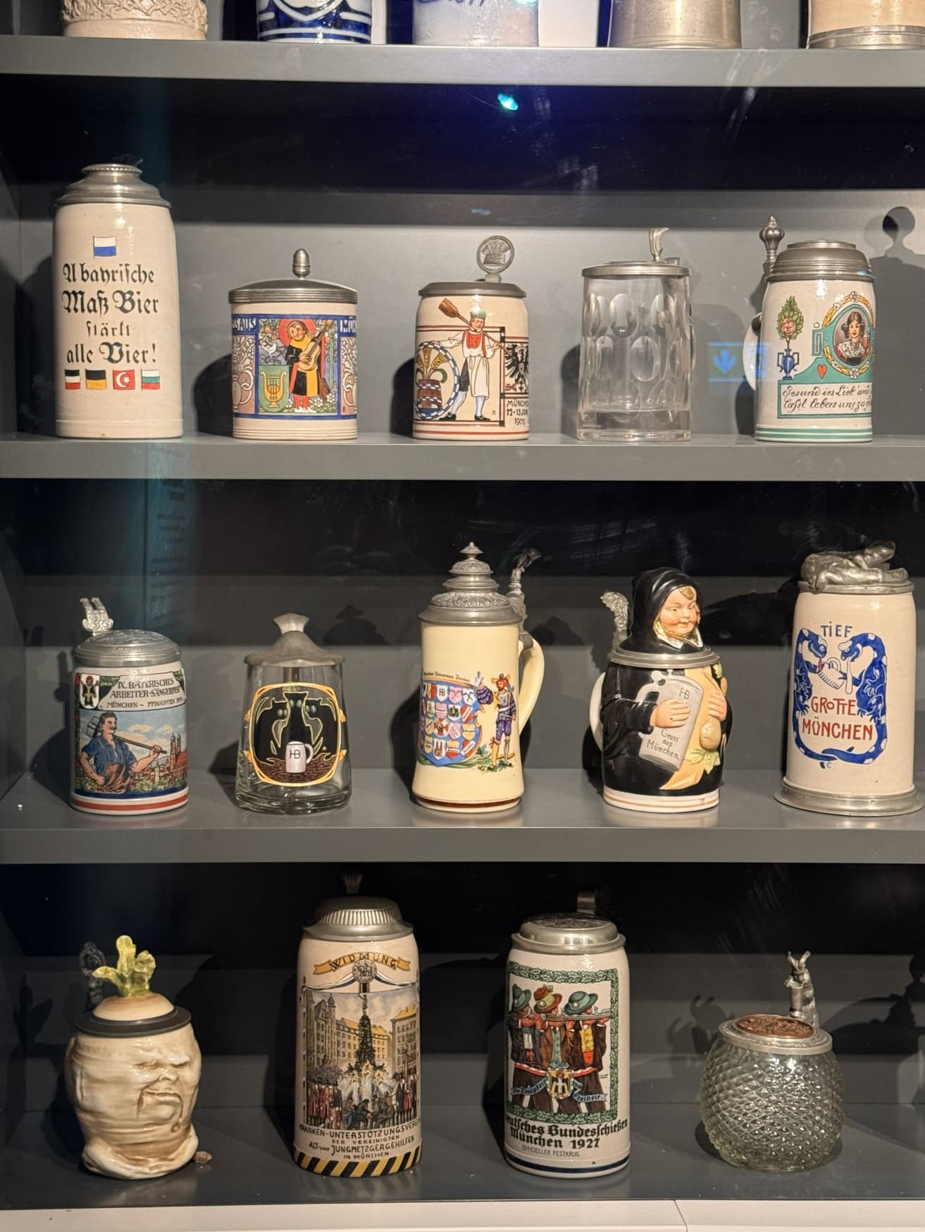 In einem dunklen Regal in einem Museum sind zahlreiche unterschiedliche Bierkrüge ausgestellt.
