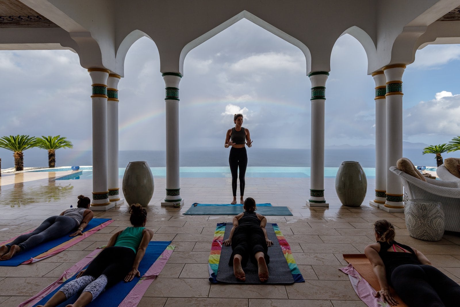 Yogastunde unter maurischen Bögen mit Blick auf das Meer und einen Regenbogen über dem Infinity-Pool.