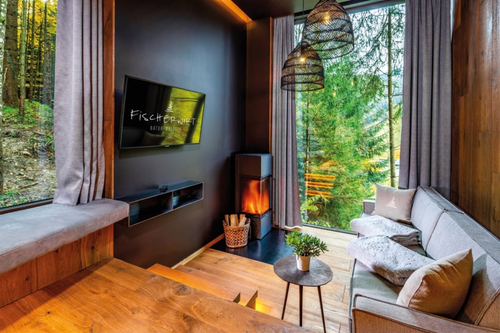 Ein kleines Hotelzimmer mit bodentiefen Fenstern und mehreren Ebenen, getrennt durch je zwei bis drei Stufen. Der Boden ist aus dunklen Holzbohlen, die Wand schwarz gestrichen. An der Wand zwischen zwei Fenstern links ein Fernseher mit der Aufschrift "Fischerwirt", gegenüber ein modernes, graues Polstersofa. In der Ecke ein hoher, moderner Kamin mit brennendem Feuer. Durch die Fenster wird der Blick auf einen dichten Wald freigegeben.