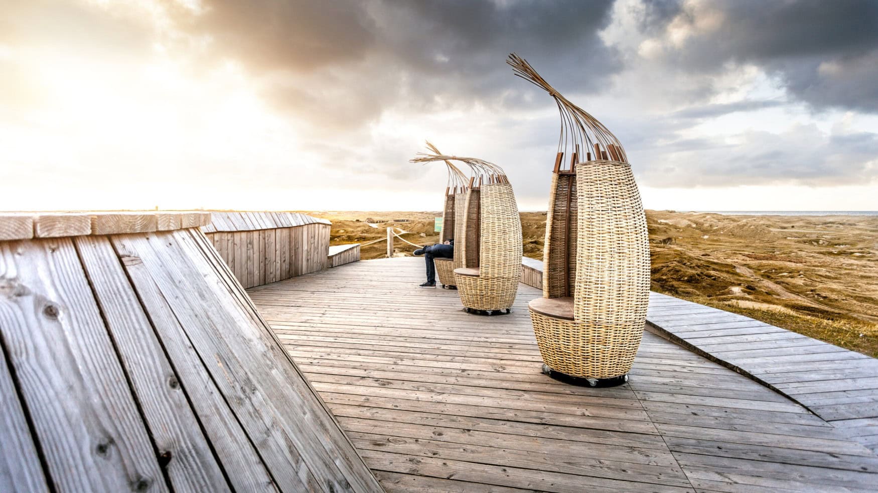 Auf einer hölzernen Plattform inmitten einiger Dünen stehen drei hohe, schmale Strandkörbe.