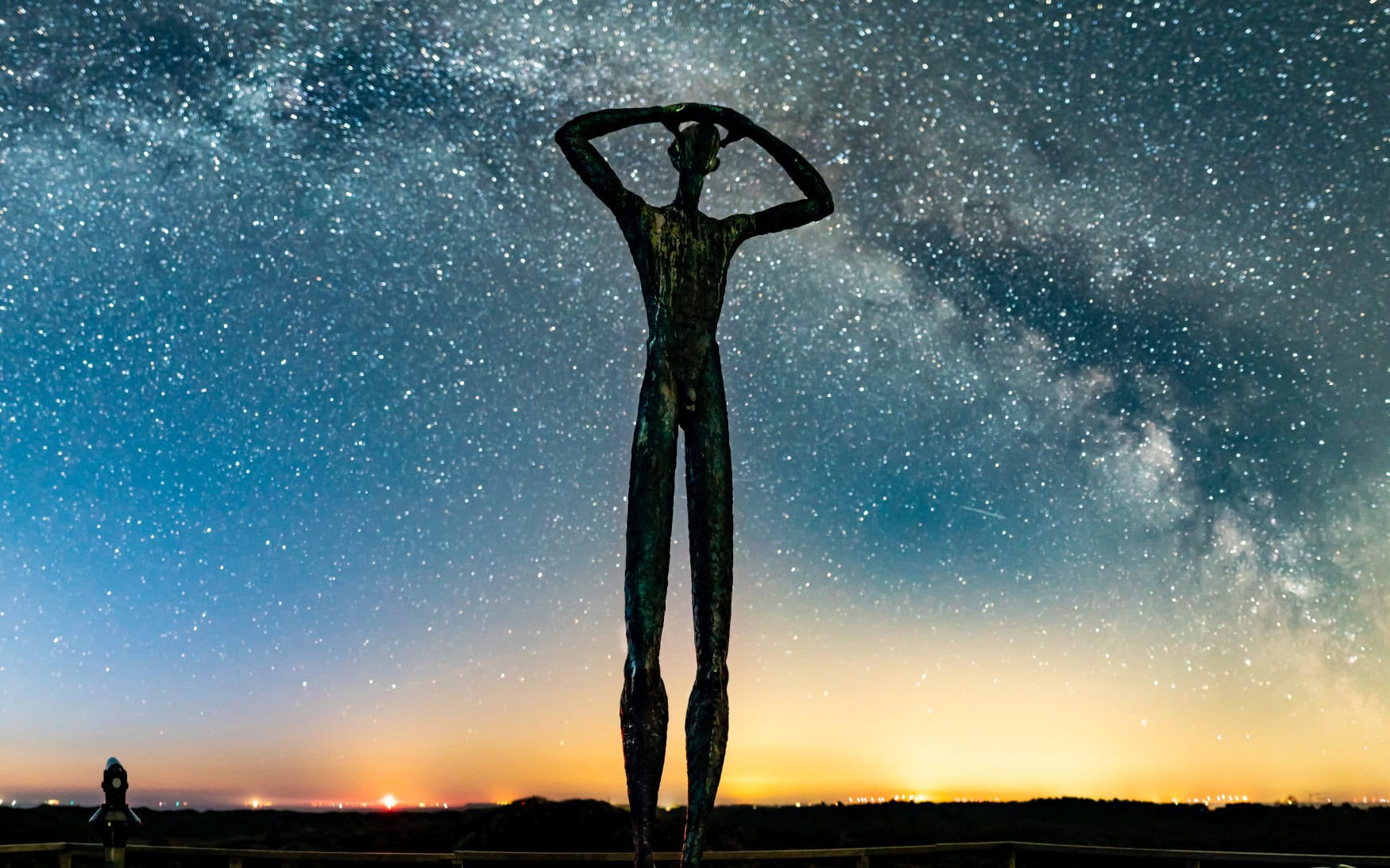 Vor einem hellen Sternenhimmel zeichnet sich die Silhouette einer Bronzefigur ab, die den Kopf gen Himmel reckt.