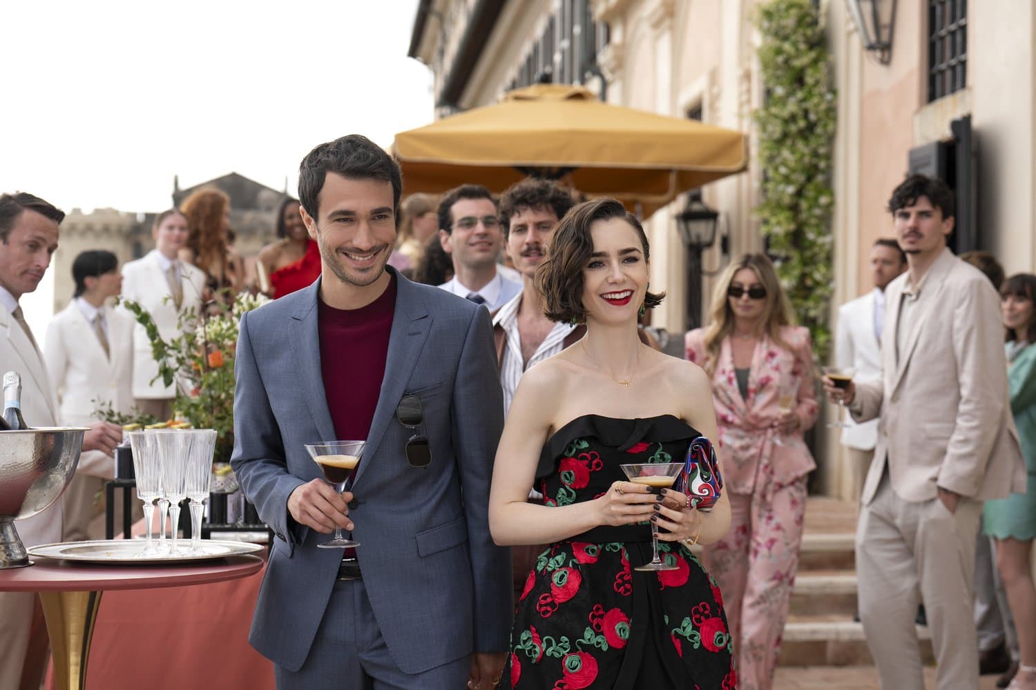 Elegantes Paar mit Espresso-Martini-Gläsern auf einer sonnigen Rooftop-Party in Italien, Frau im schwarzen Kleid mit roten Rosen, Mann im hellblauen Anzug, weitere Gäste im Hintergrund