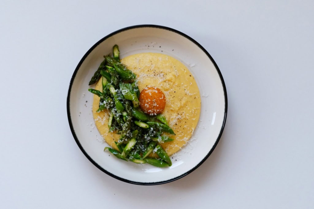 In einem tiefen Teller ist eine Portion Polenta mit gehacktem, grünen Spargel angerichtet. Genau in der Mitte ist ein pochiertes Eigelb platziert, über alles wurde Käse gerieben.