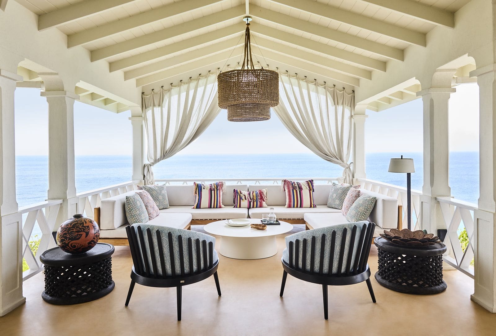 Offener Pavillon mit weißem Sofa, schwarzen Designersesseln und gerafften Vorhängen vor dem Meer.