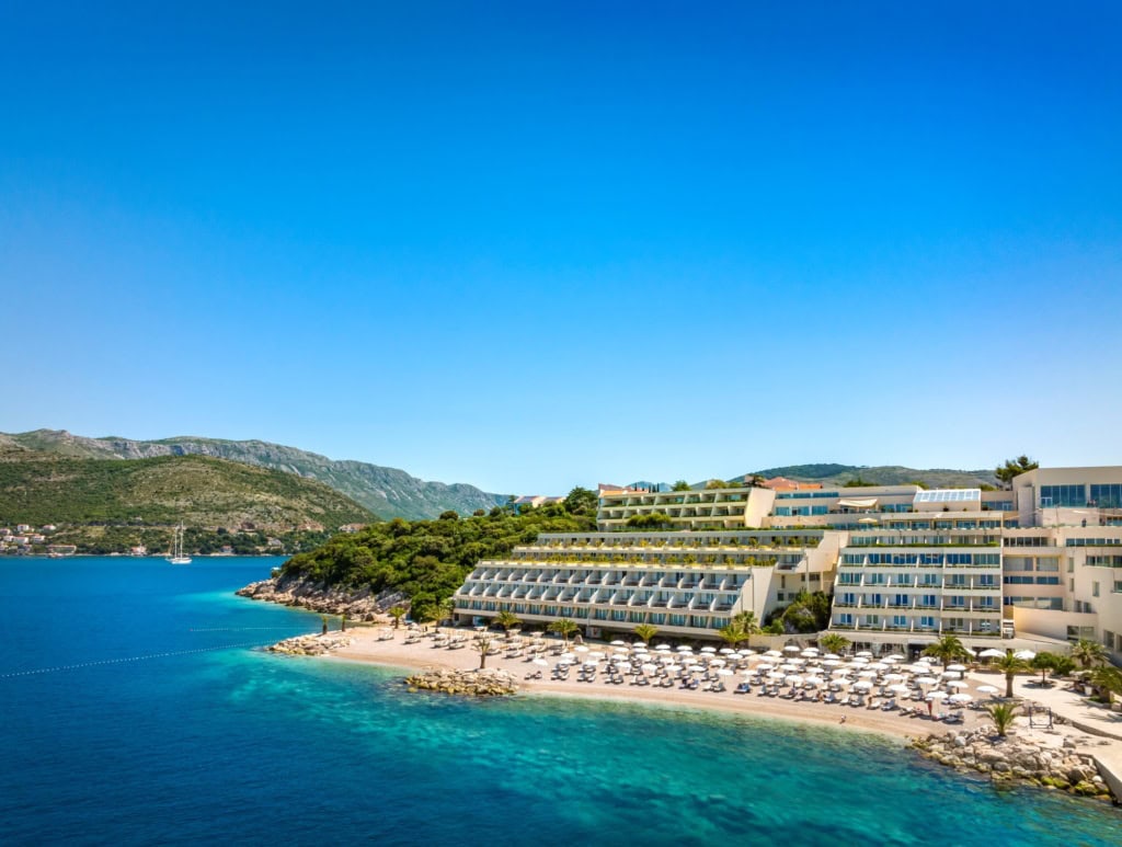 Ein Strand in Kroatien unter blauem Himmel. Zur rechten liegt ein großer, weißer Hotelkomplex im Licht der Mittagssonne.