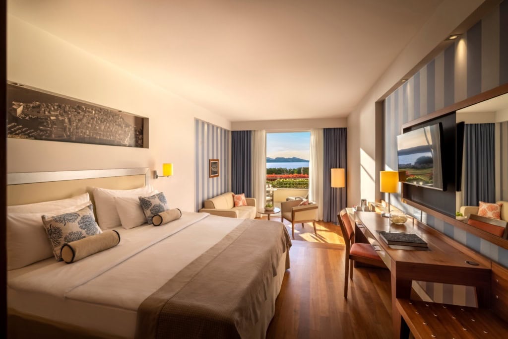Ein geräumiges Zimmer im President Hotel, Valamar Collection in Kroatien. Links ein breites Doppelbett, rechts ein Schreibtisch mit Fernseher darüber, im Hintergrund eine offene Balkontür, die den Blick auf die Adria freigibt.