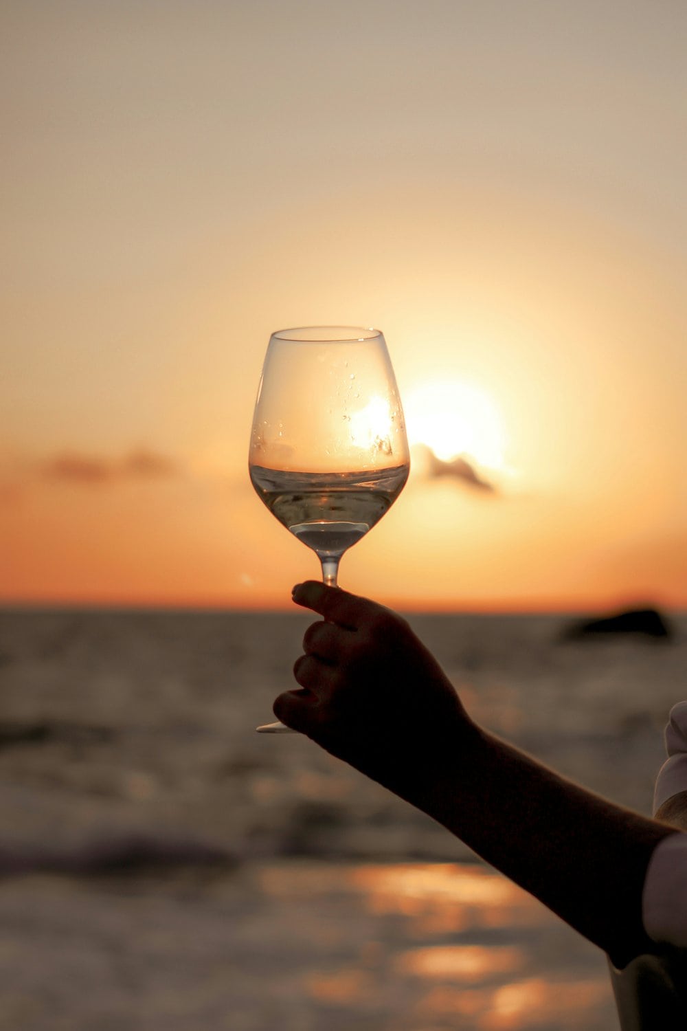 Hand hält ein Weißweinglas in die Abendsonne, im Hintergrund das Meer und der Horizont in warmem Orange.