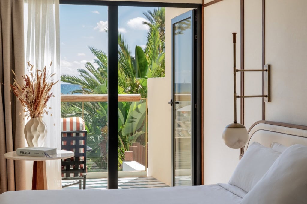 Bless Ibiza The Site - Premium Raum, im Vordergrund ein Bett mit glatten weißen Laken, links ein eleganter Beistelltisch mit Buch und Trockenblumen, die Balkontür ist geöffnet und zeigt das Meer, Palmen.