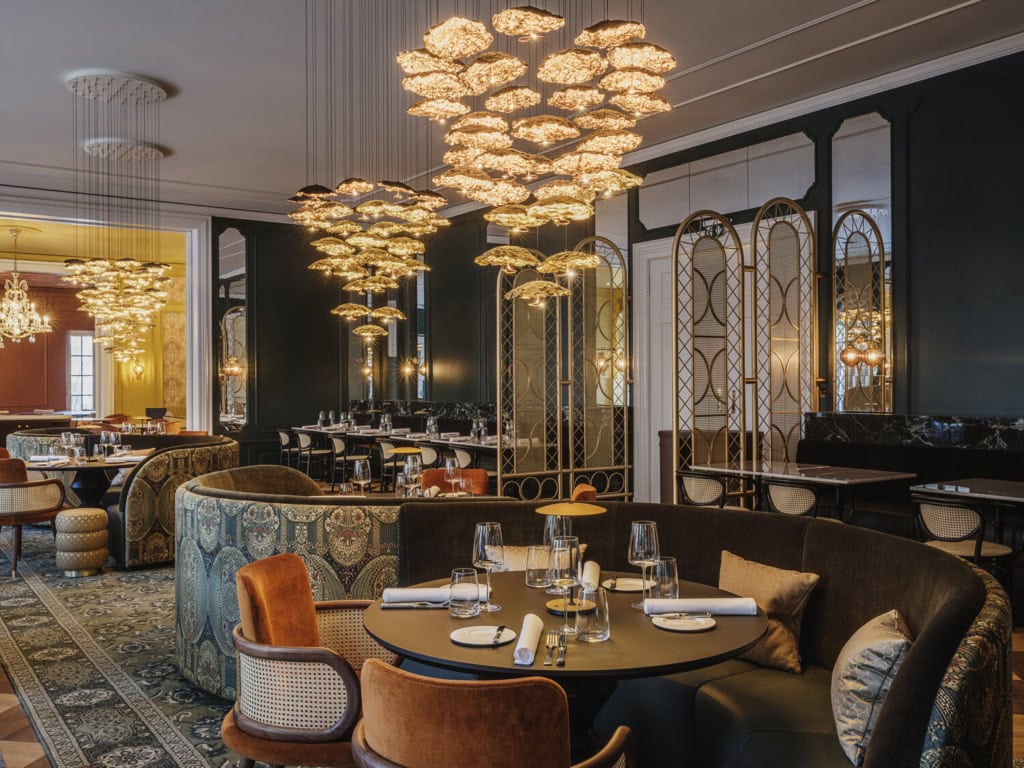 Elegantes Restaurant mit dunkelgrünen Wänden, opulenten goldenen Blüten-Kronleuchtern, gemusterten Polstersofas, samtbezogenen Sesseln und gedeckten Tischen auf orientalischem Teppich.