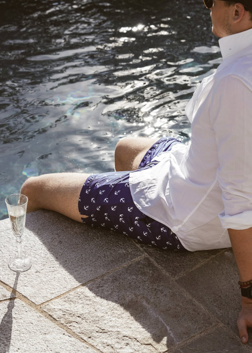 Mann in Badehose mit Ankermuster und weissem Hemd sitzt mit Champagnerglas am sonnigen Beckenrand.