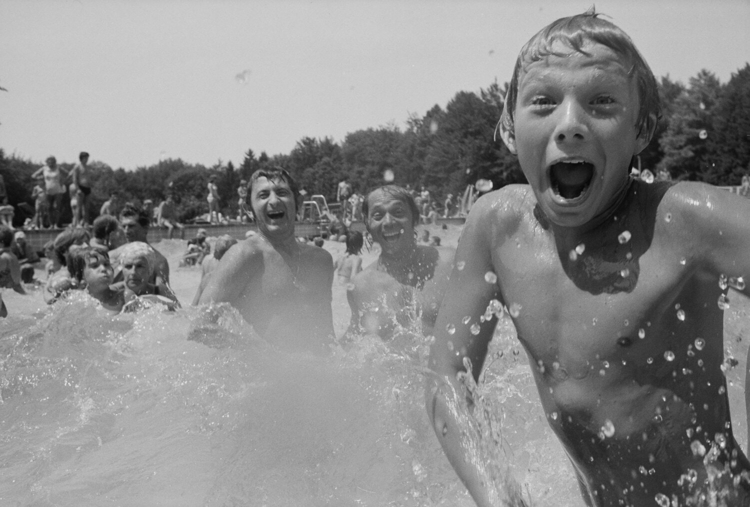 Schwarzweiss-Aufnahme: Lachender Junge spritzt im voll besetzten Freibad Wasser, zahlreiche Badegaeste im Hintergrund.