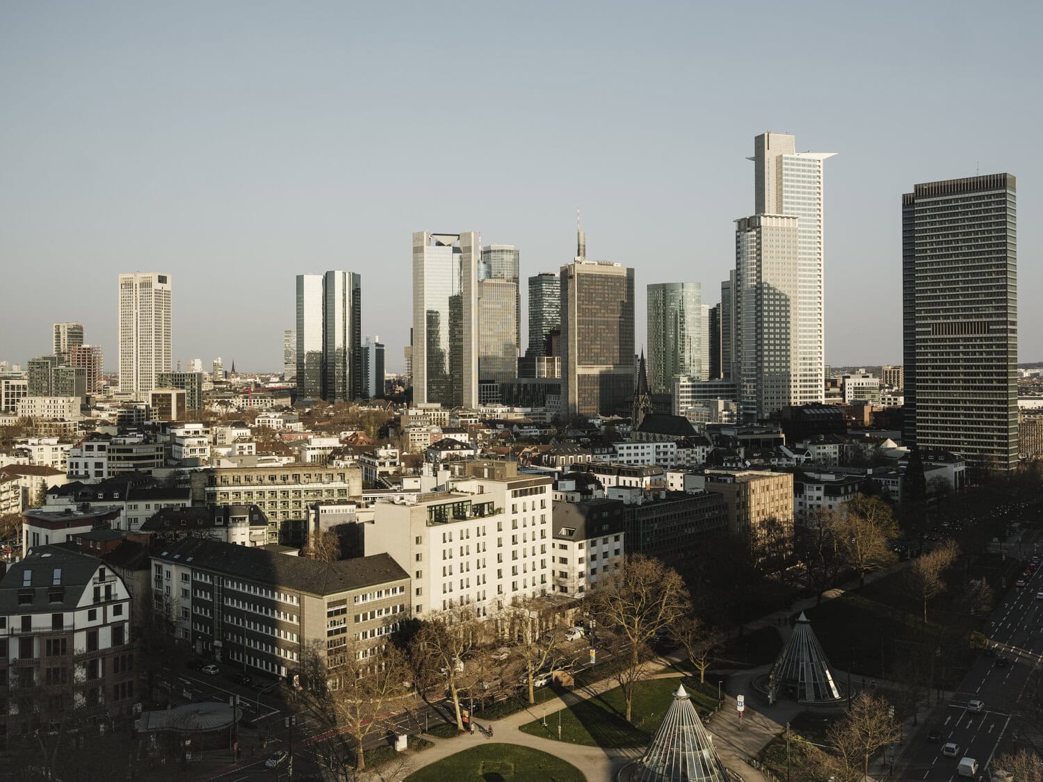 Blick über die Frankfurter Skyline mit Hochhäusern des Bankenviertels bei Tageslicht, im Vordergrund niedrigere Wohngebäude und eine begrünte Parkanlage.