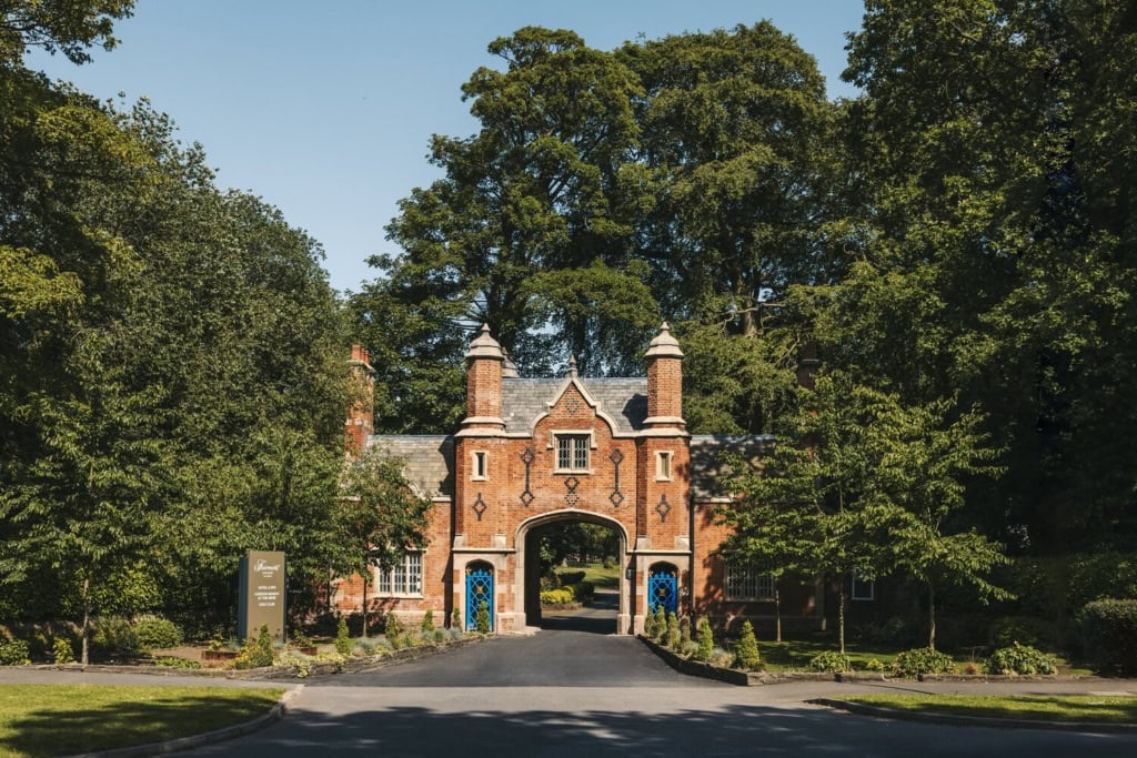 Historisches Torhaus aus rotem Backstein im Tudorstil mit spitzem Mittelbogen, eingerahmt von hohen grünen Bäumen, am Ende einer Zufahrtsstraße bei Sonnenschein.
