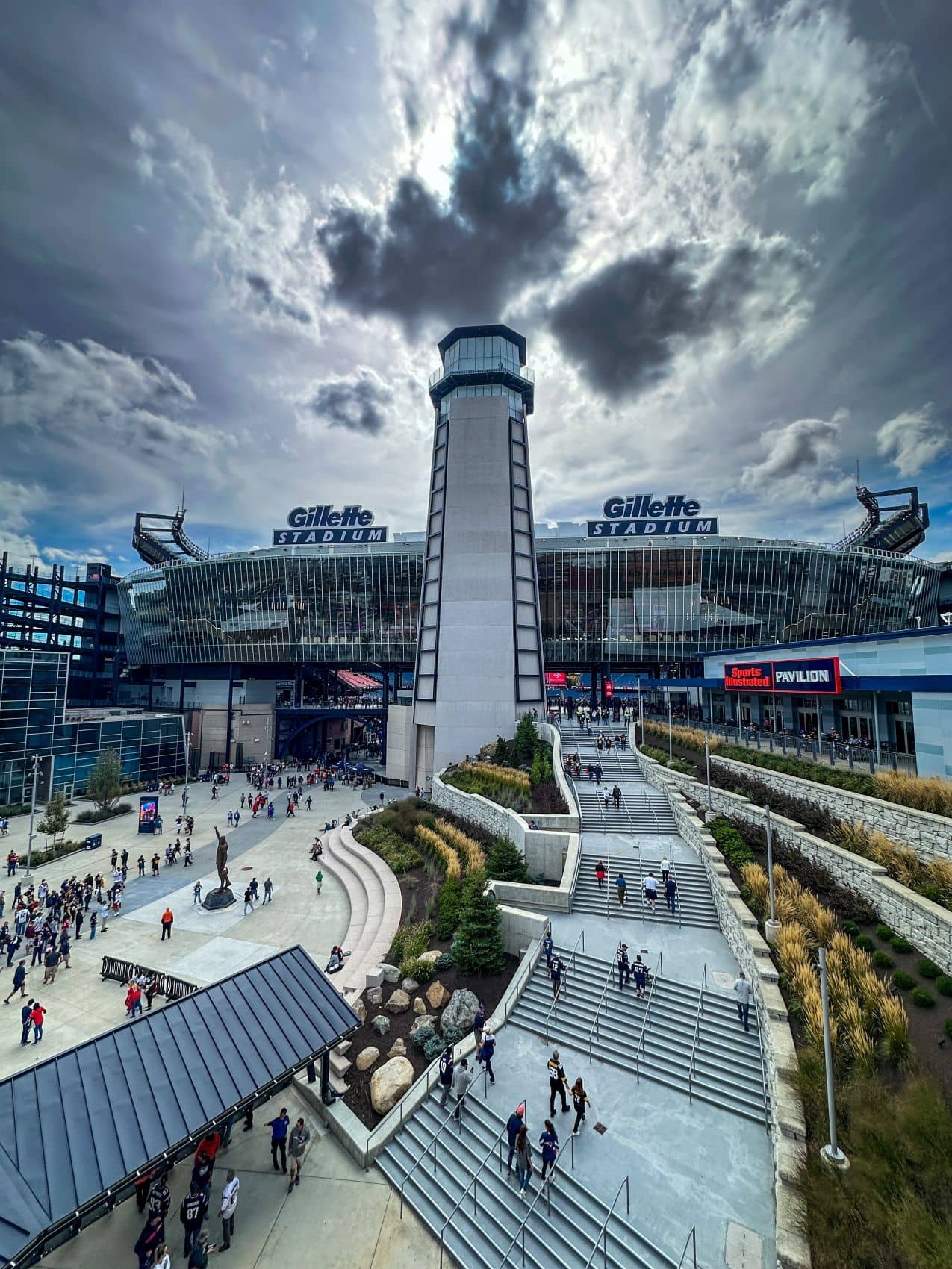 Besucher strömen über Treppen und Wege zum Gillette Stadium in Foxborough bei Boston. Über dem markanten Aussichtsturm ziehen dunkle Wolken auf.