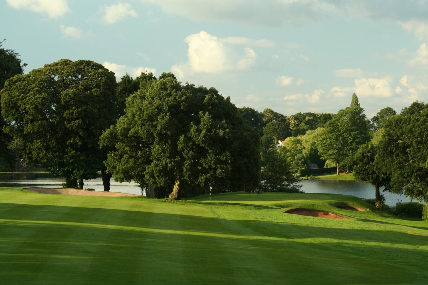 Gepflegtes Golfgrün mit Fahne und Bunkern, umgeben von alten Laubbäumen und einem See unter blauem Himmel mit leichten Wolken.