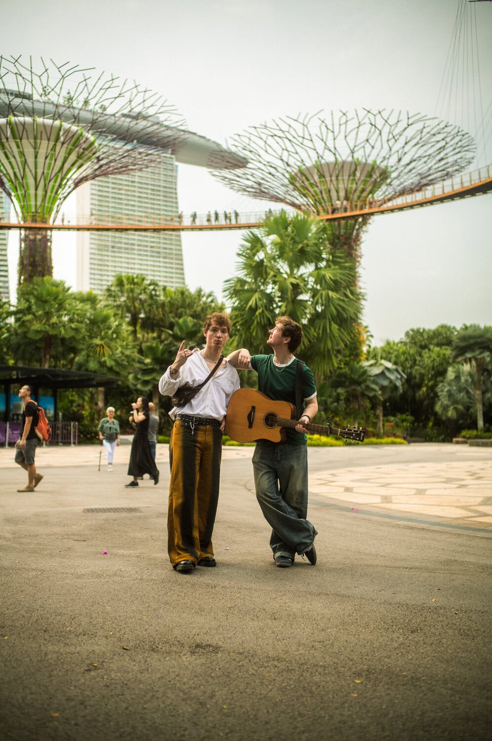 Heiko und Roman Lochmann gehen mit Akustikgitarre über einen Platz vor den Supertrees der Gardens by the Bay in Singapur.