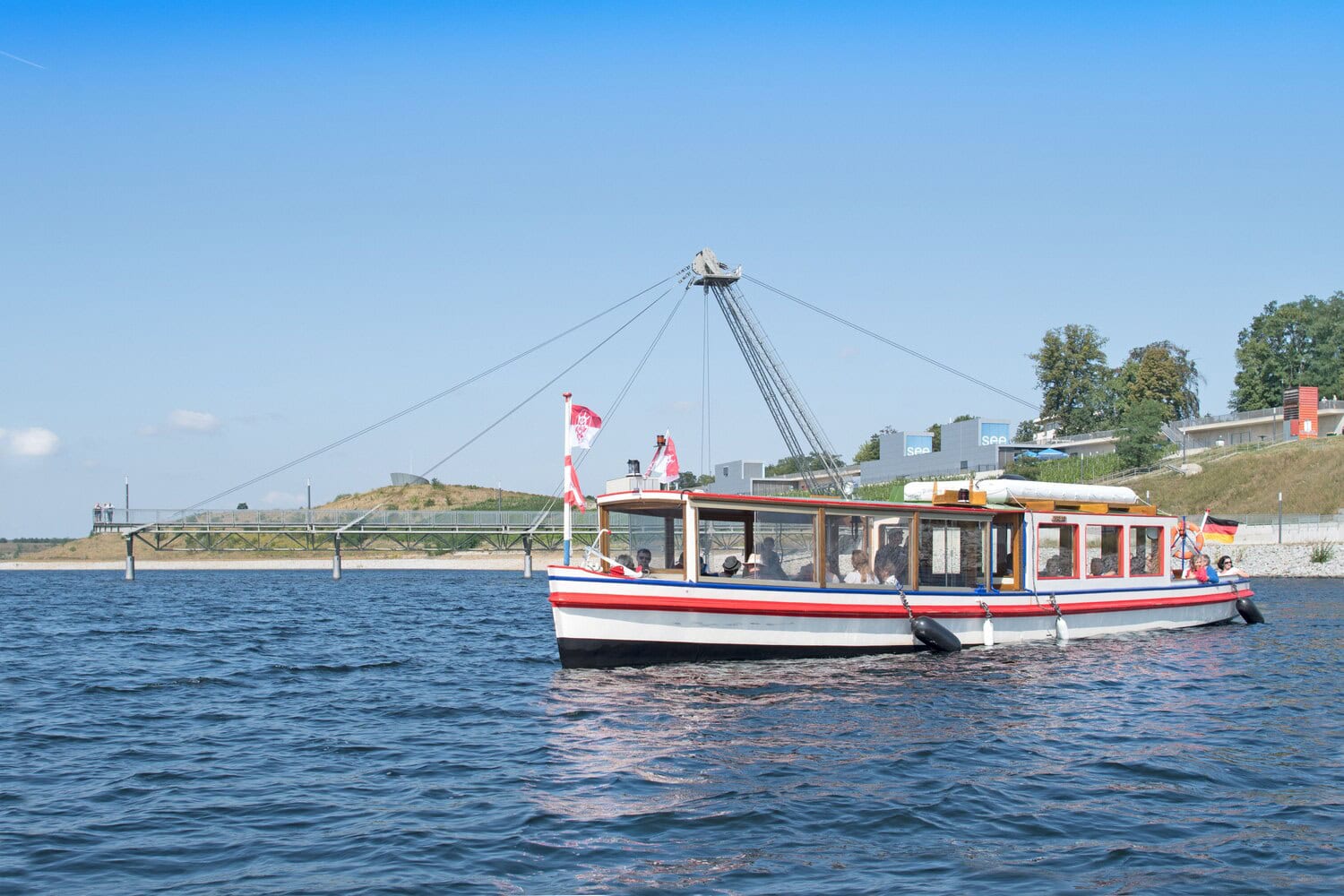 Kleines Fahrgastschiff mit Flaggen auf dem Grossraeschener See, im Hintergrund eine Seebruecke.