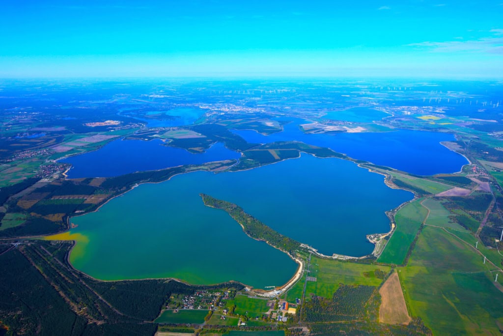 Weite Luftaufnahme des Lausitzer Seenlands mit mehreren Seen, Feldern und Windraedern am Horizont.