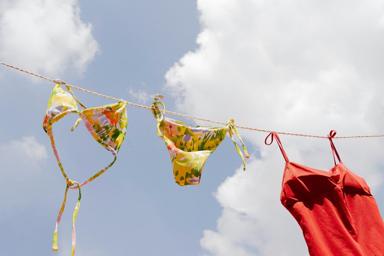 Bunter Bikini und rotes Kleid haengen an einer Waescheleine vor blauem Himmel mit Wolken.