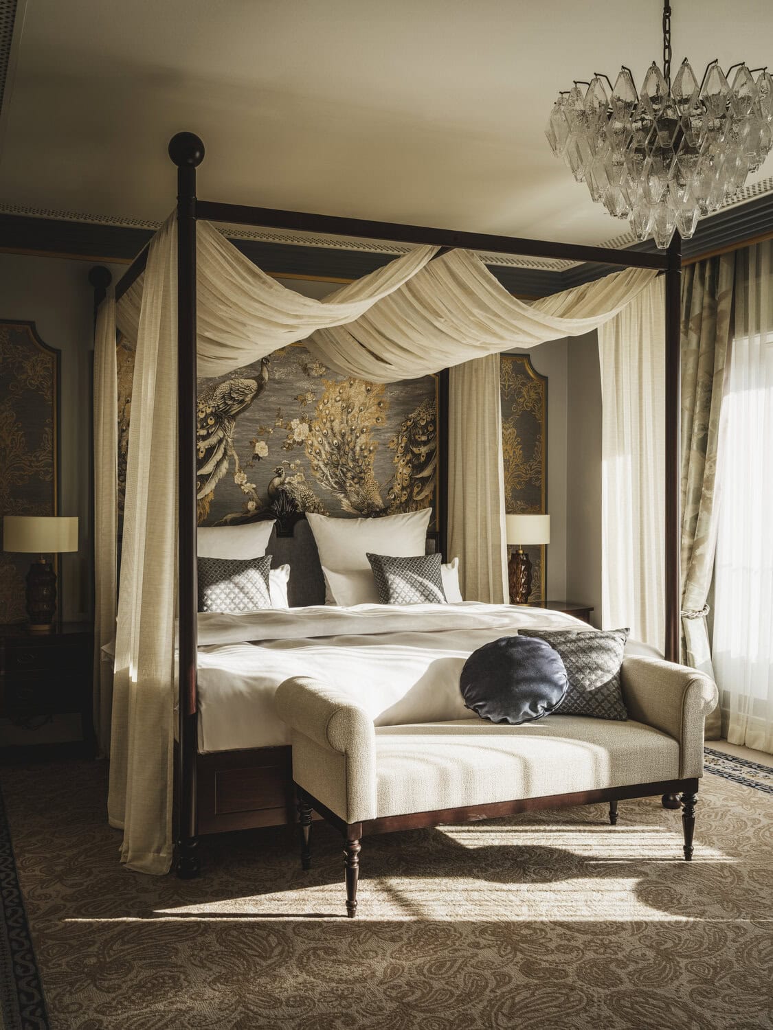 Luxuriöses Schlafzimmer einer Suite mit dunklem Himmelbett samt cremefarbenen Vorhängen, kunstvollem Wandbild hinterm Bett, Glas-Kronleuchter und gepolsterter Sitzbank am Fußende.Suite im Taj Hessischer Hof