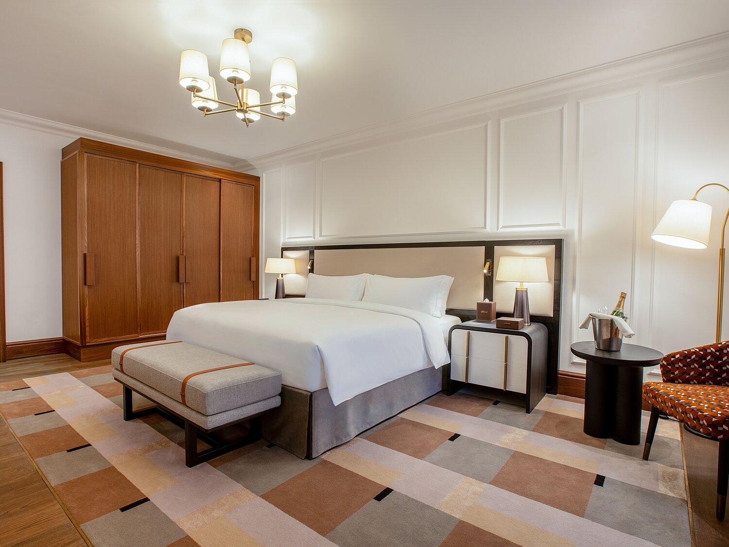 Geräumiges Suiten-Schlafzimmer mit Kingsize-Bett, Holzkleiderschrank, Sessel und Champagnerkühler, kariertem Teppich in Erdtönen und weißer Wandvertäfelung.
